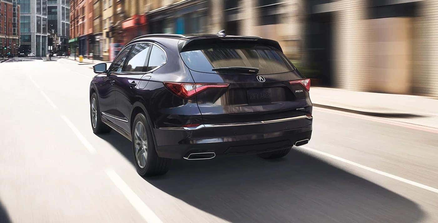 Black 2023 Acura MDX driving on the road with buildings in the background.