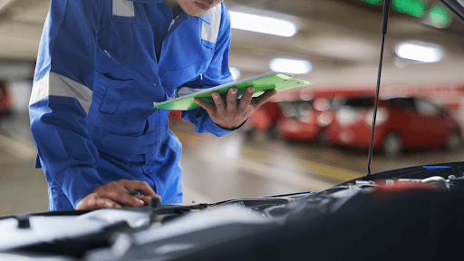 A mechanic holding a clipboard and inspecting the hood of a car. 
