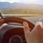 driver hands on steering wheel with open highway woods and mountains ahead