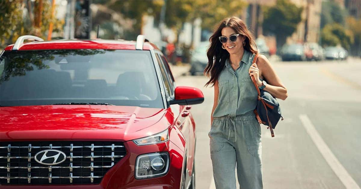 A smiling woman in a green jumpsuit and sunglasses walks past a red 2026 Hyundai Venue parked on a city street.