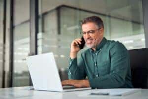 An older middle aged man smiling while making a phone call on a cell phone and using laptop computer sitting at desk in an office