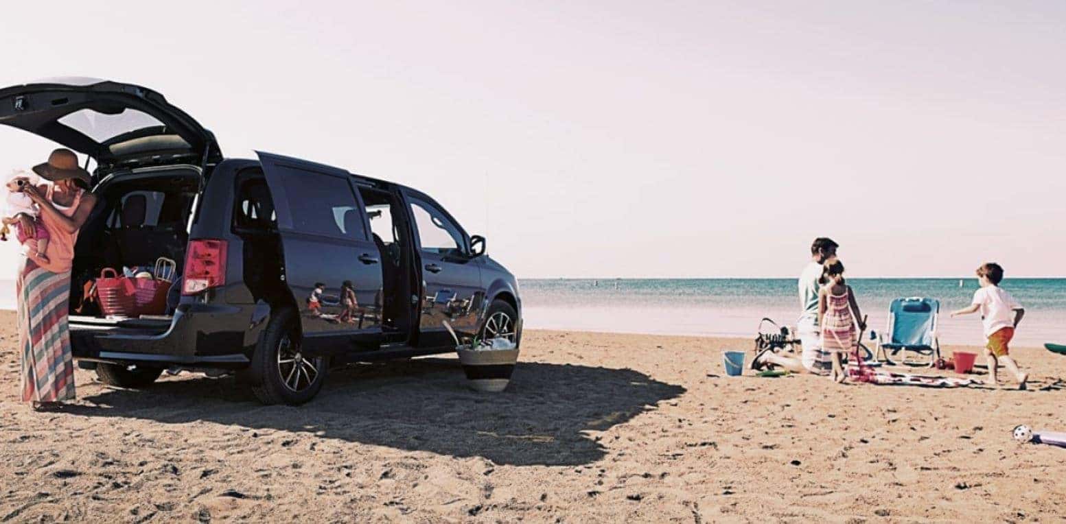 family at the bach with their dodge grand caravan on the sand while mom holds infant and dad is playing with toddler aged son and daughter