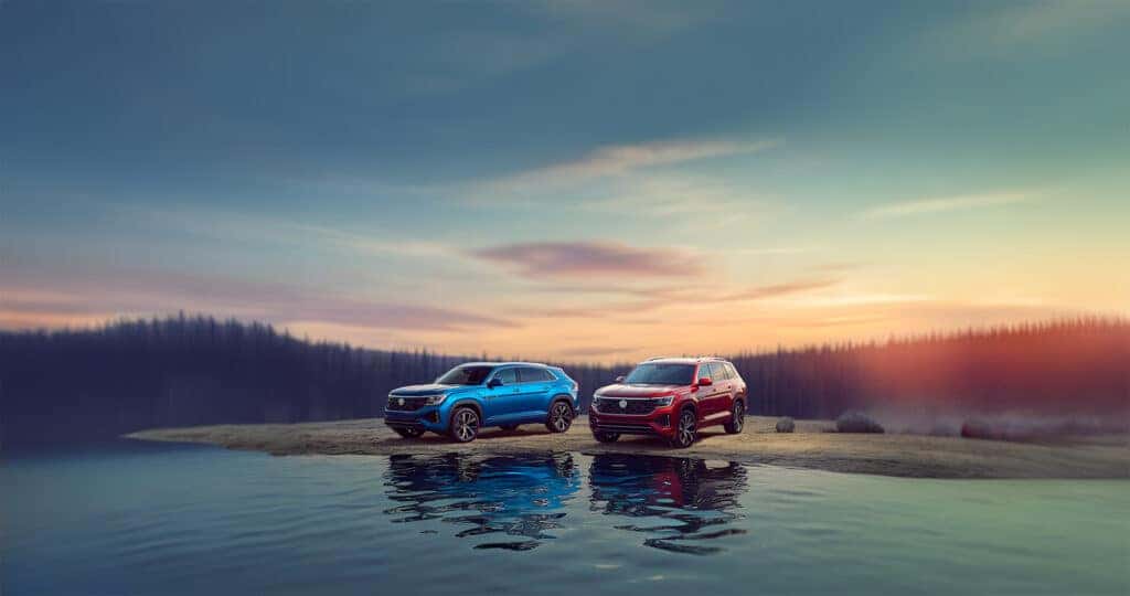 2026 Volkswagen Atlas vs Atlas Cross Sport parked next to eachother at the edge of a mountain lake at sunset