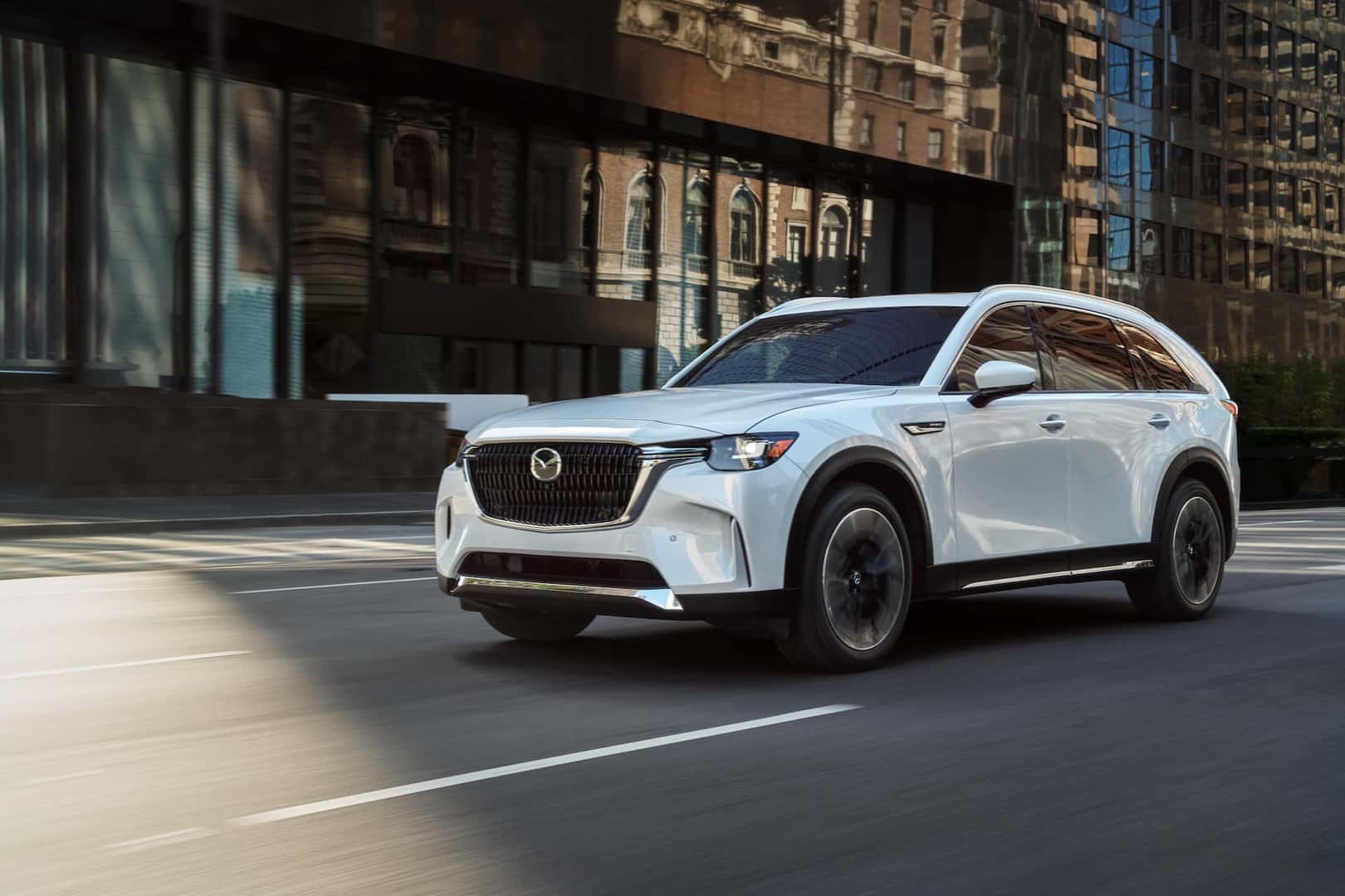 White Mazda CX-90 PHEV driving on a road in a city.