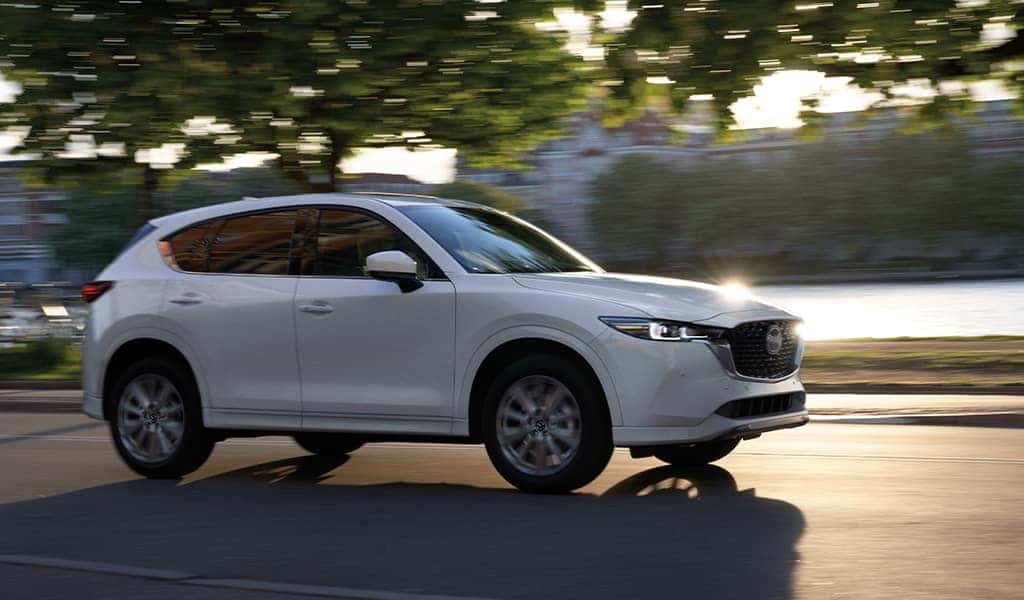 A white 2023 Mazda CX-5 vehicle driving on the road with a lake and buildings in the background.