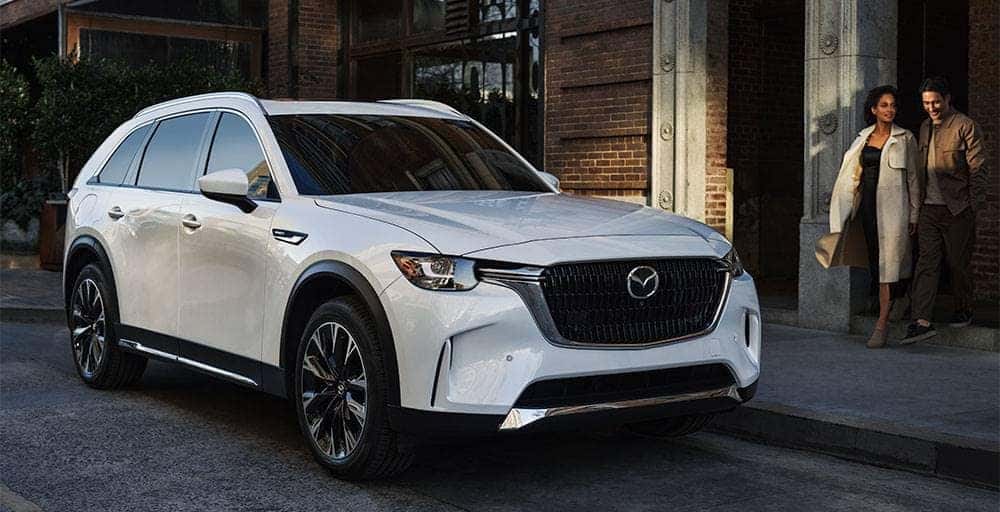 White 2024 Mazda CX-90 parked on the side of a city street with two individuals walking towards it.