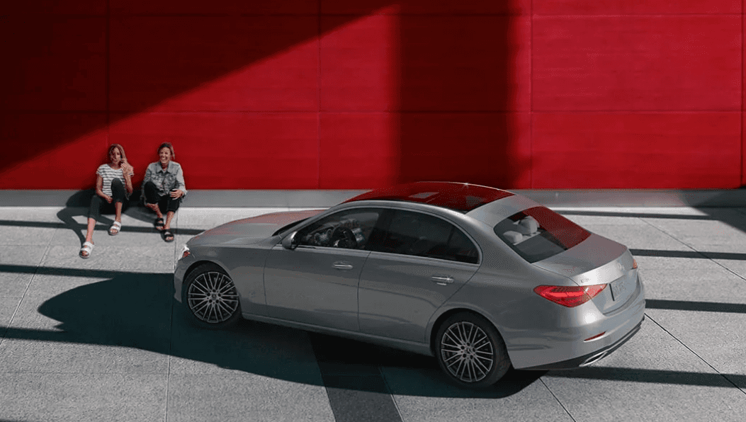 A parked 2024 silver Mercedes-Benz. Long shadows are cast over it and on the pavement and two women sit against a red wall behind it