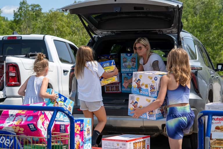 Three children carrying cases of snacks to a waiting SUV while a woman loads the back