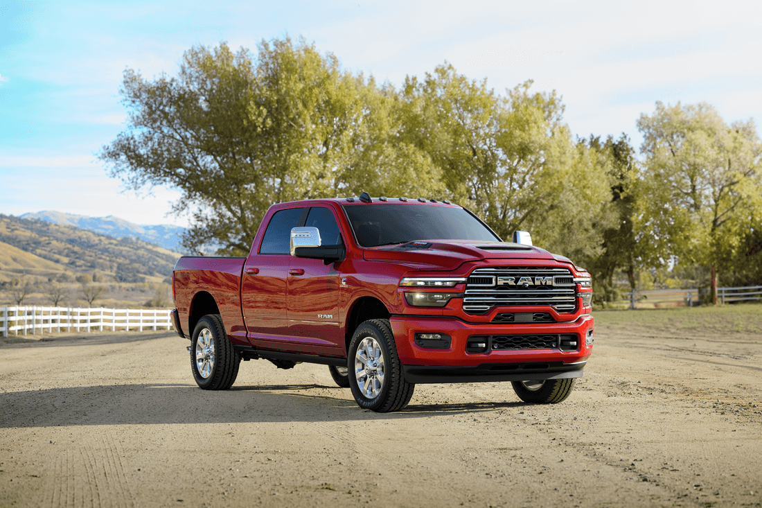 2025 2500 Laramie HD with Sport Appearance Package in Red Hot parked in a fenced dirt lot, desert mountains, trees and cloudy blue sky visible behind.