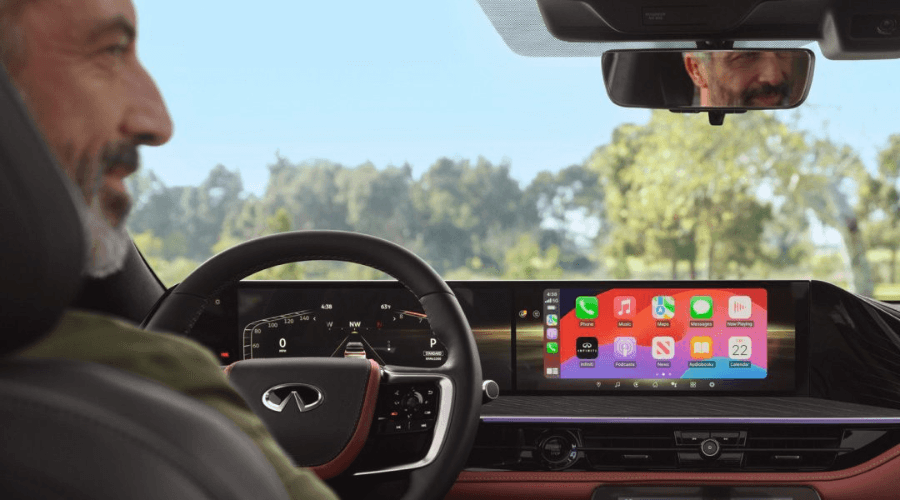 Interior view of the 2026 INFINITI QX80 front cabin from behind the driver's seat, highlighting the dual-screen digital dashboard displaying Apple CarPlay, a leather-wrapped steering wheel, and premium burgundy leather interior accents at Ramsey INFINITI in Ramsey, NJ