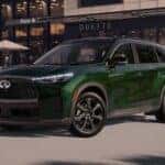 Front three-quarter view of a 2026 INFINITI QX60 3-row luxury SUV in Deep Emerald green paint with an illuminated front grille, parked outside a boutique shop in Ramsey, NJ.