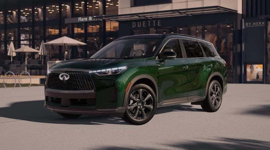 Front three-quarter view of a 2026 INFINITI QX60 3-row luxury SUV in Deep Emerald green paint with an illuminated front grille, parked outside a boutique shop in Ramsey, NJ.
