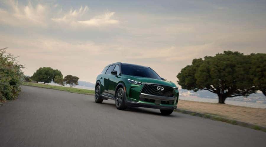 Front three-quarter view of a 2026 INFINITI QX60 3-row luxury SUV in Deep Emerald green driving along a coastal road near Mahwah and Ramsey, NJ.