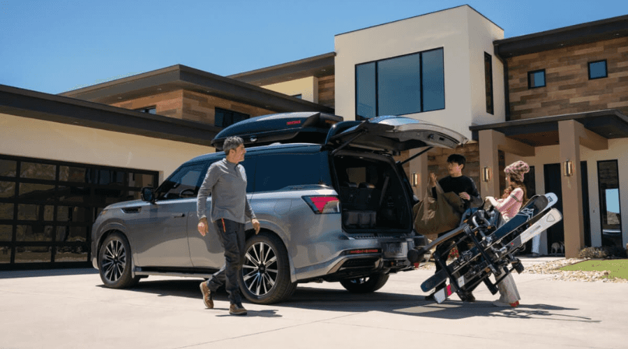 Family loading luggage, skis, and a Yakima roof cargo box into the open tailgate of a silver 2026 INFINITI QX80 parked outside a luxury Bergen County home before a road trip from Ramsey NJ
