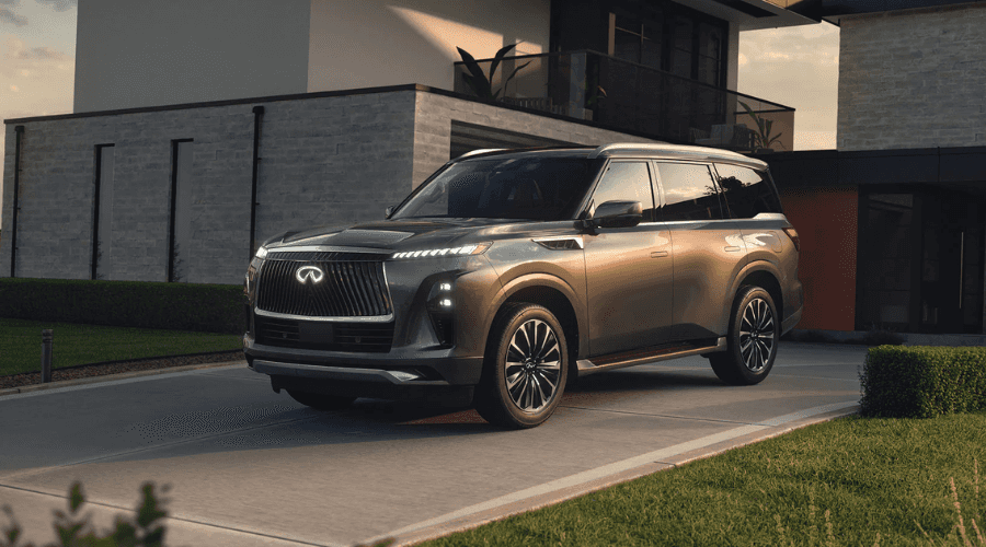 Front three-quarter view of a dark metallic 2026 INFINITI QX80 luxury SUV with illuminated grille badge parked in the driveway of a modern Bergen County home near Ramsey NJ