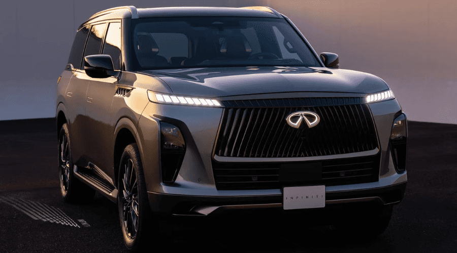 Front view of a dark metallic 2026 INFINITI QX80 luxury 3-row SUV displaying bright illuminated LED headlights and double-arch grille outside a garage in Ramsey, NJ, featuring Biometric Cooling climate control technology.
