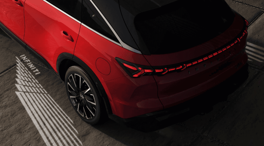 Rear three-quarter overhead view of a red 2026 INFINITI QX65 displaying full-width LED taillights and welcome puddle lighting projecting the INFINITI logo carpet onto the pavement at Ramsey INFINITI in Bergen County, NJ.