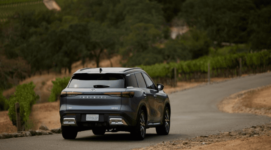 Rear three-quarter view of a blue 2026 INFINITI QX60 luxury SUV driving along a scenic winding road, showcasing its full-width LED taillights and rear camera sensor array available at Ramsey INFINITI in Bergen County, NJ.