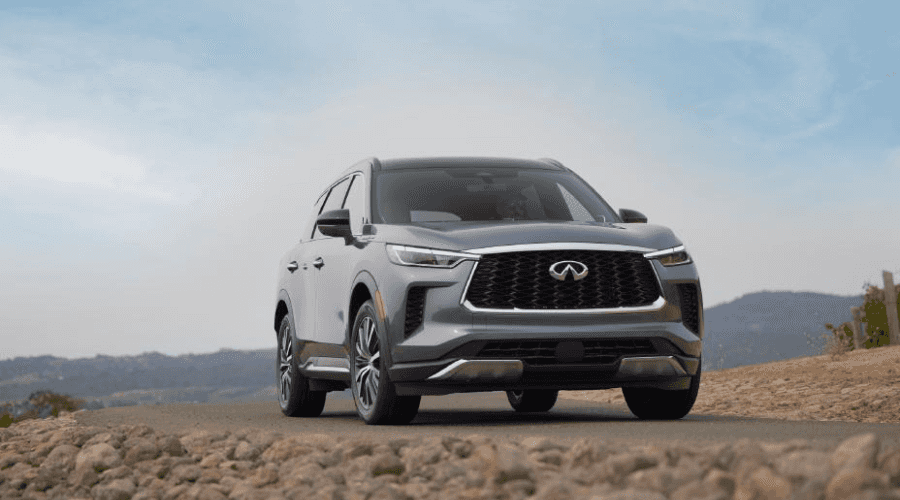 Low-angle front view of a metallic grey 2026 INFINITI QX60 luxury SUV driving on a paved road, showcasing the signature double-arch grille, LED headlights, and front camera sensor housing available at Ramsey INFINITI in Bergen County, NJ.