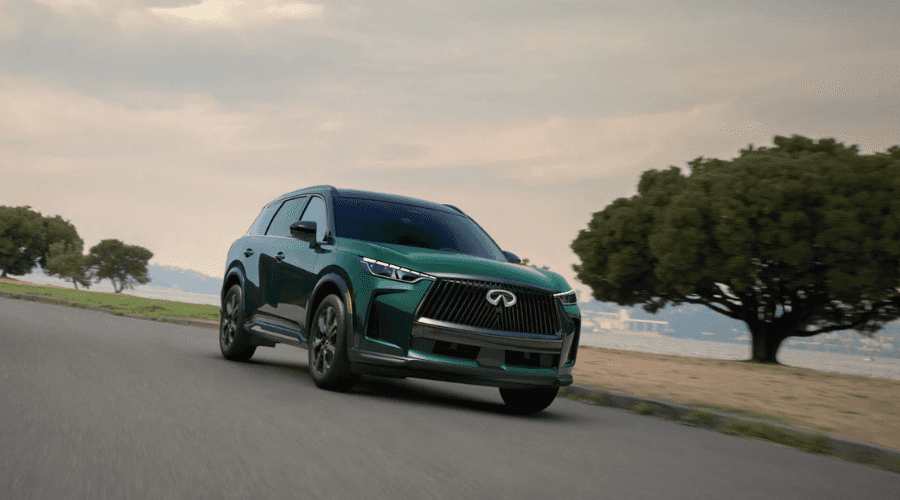 Deep green 2026 INFINITI QX60 luxury SUV with Intelligent AWD navigating a curved road under overcast skies near Ramsey, NJ.