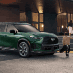 2027 INFINITI QX60 Luxury 3-Row SUV in Deep Emerald Green parked in front of a modern performance arts venue as a family with children walk towards the entrance, available at Ramsey INFINITI serving Bergen County, NJ.