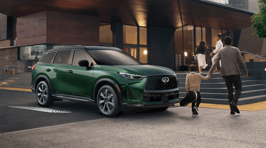 2027 INFINITI QX60 Luxury 3-Row SUV in Deep Emerald Green parked in front of a modern performance arts venue as a family with children walk towards the entrance, available at Ramsey INFINITI serving Bergen County, NJ.