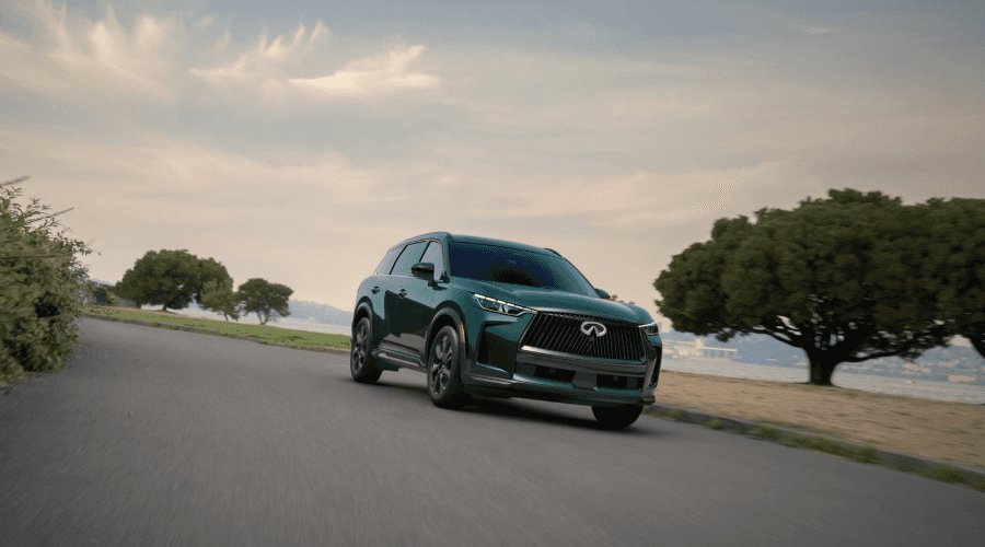 Front three-quarter view of a deep emerald green 2026 INFINITI QX60 driving along a scenic coastal road, showcasing its signature double-arch grille, LED headlights, and sleek profile available at Ramsey INFINITI in New Jersey.