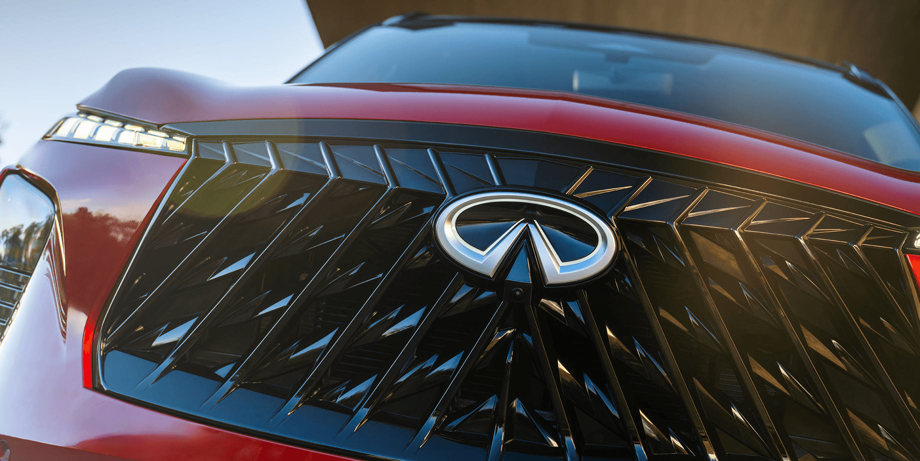 Close-up of the 2027 INFINITI QX65 front grille with illuminated emblem, black geometric mesh pattern, and slim LED lighting.