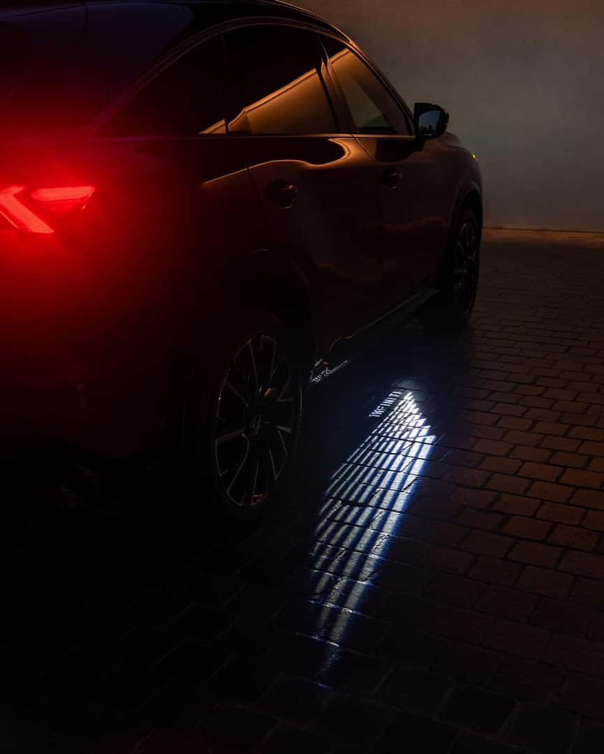 Rear side view of the 2027 INFINITI QX65 at night, showing illuminated taillight styling and welcome lighting projected onto the pavement.