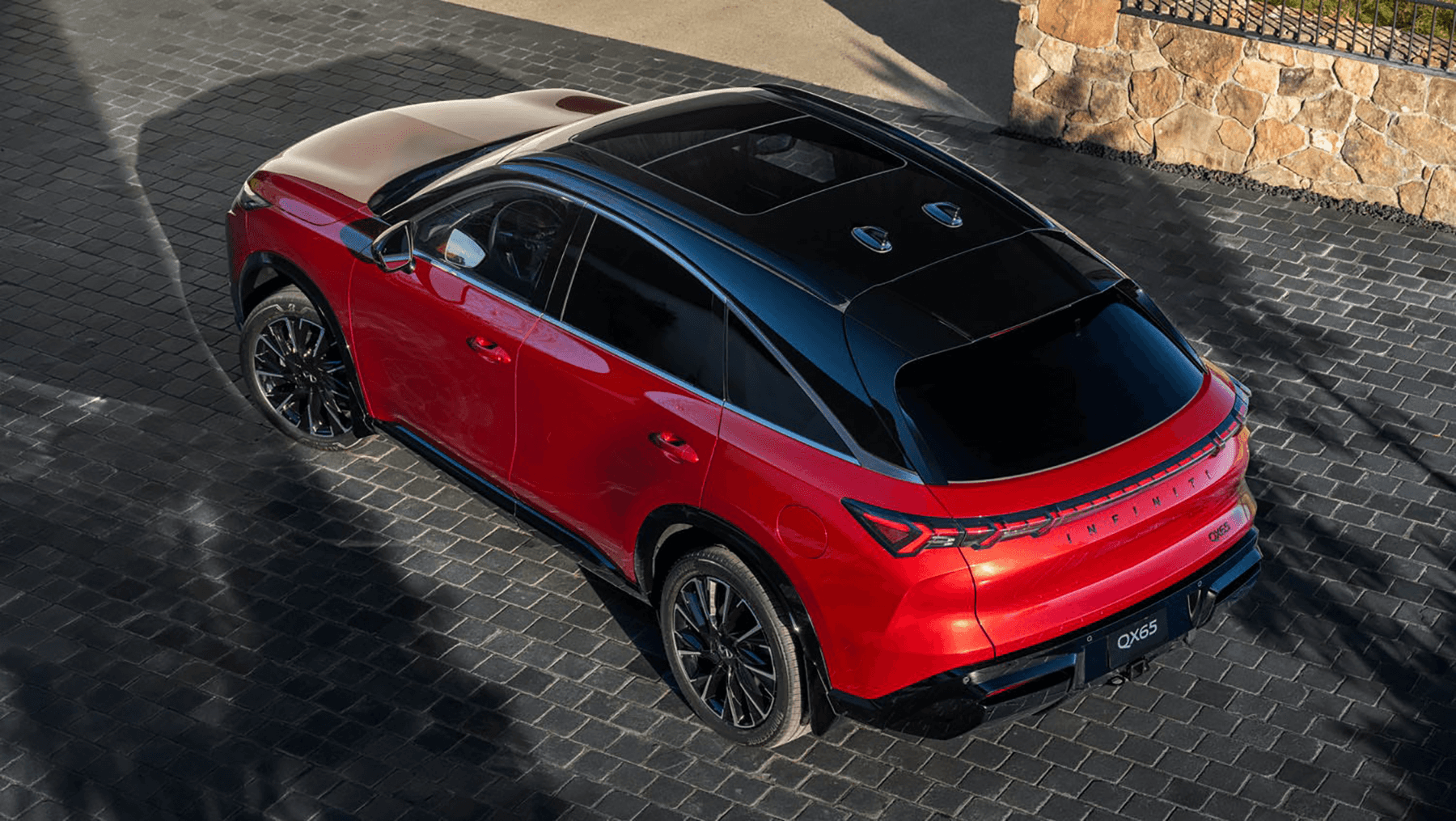 Overhead rear three-quarter view of the 2027 INFINITI QX65 in red with a contrasting black roof and panoramic glass roof.