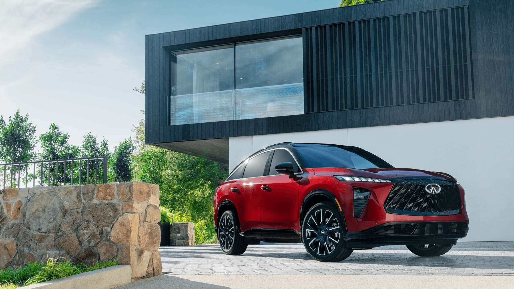 Front three-quarter view of the 2027 INFINITI QX65 parked outside a modern home, featuring red paint, black roof, and bold grille.