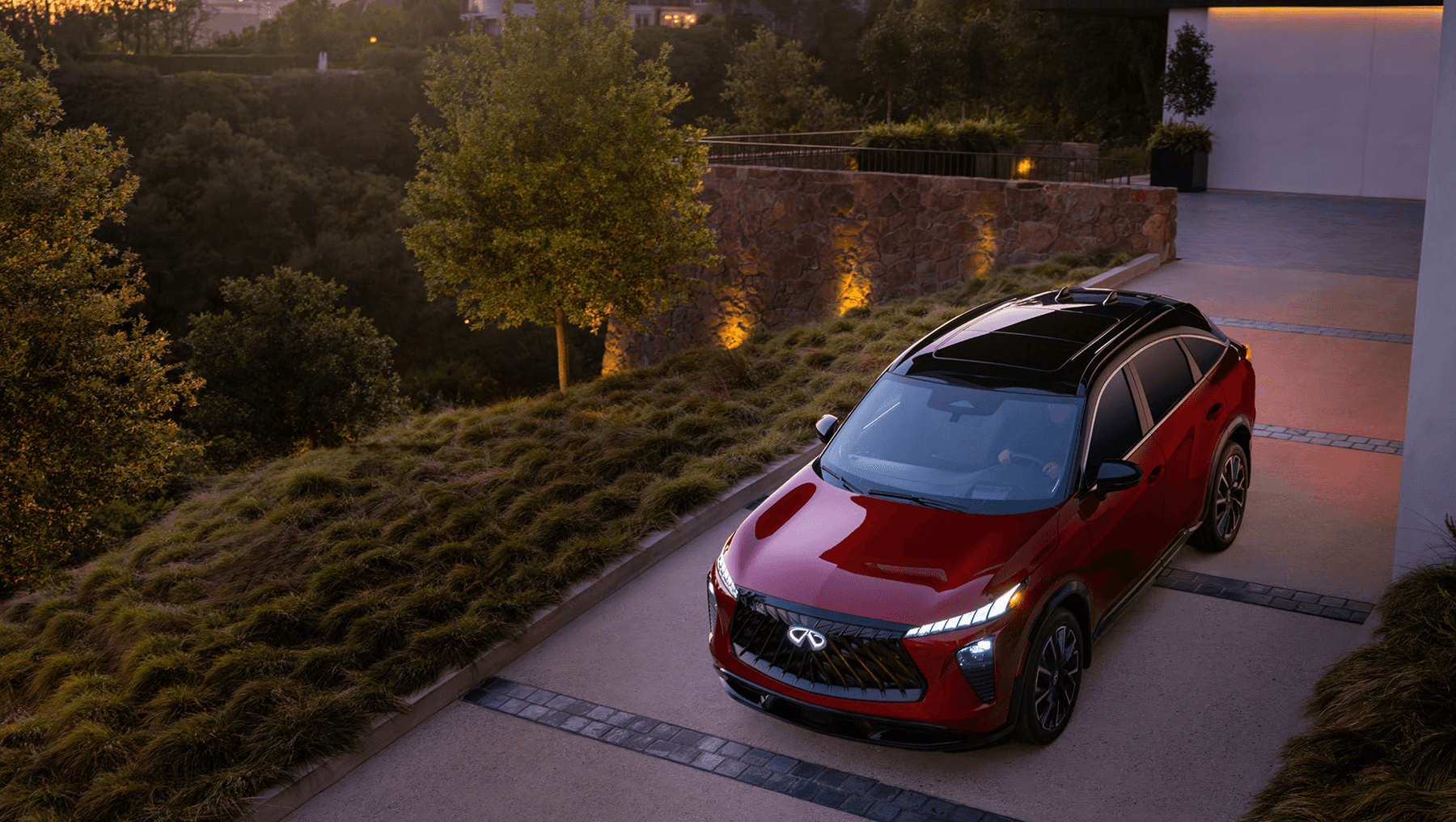 High-angle view of the 2027 INFINITI QX65 parked on a driveway at dusk, showing the black roof, panoramic glass, and illuminated front lights.