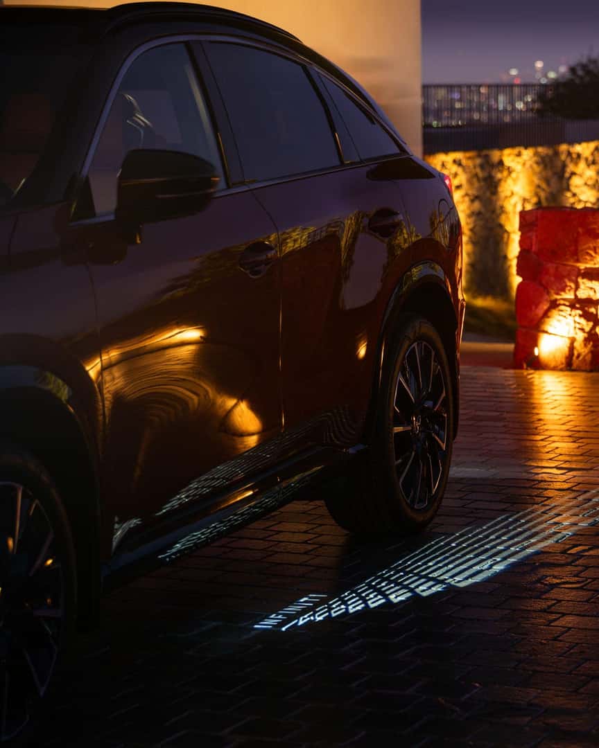 Side view of the 2027 INFINITI QX65 at dusk, highlighting glossy red paint, dark window trim, and welcome lighting on the ground.