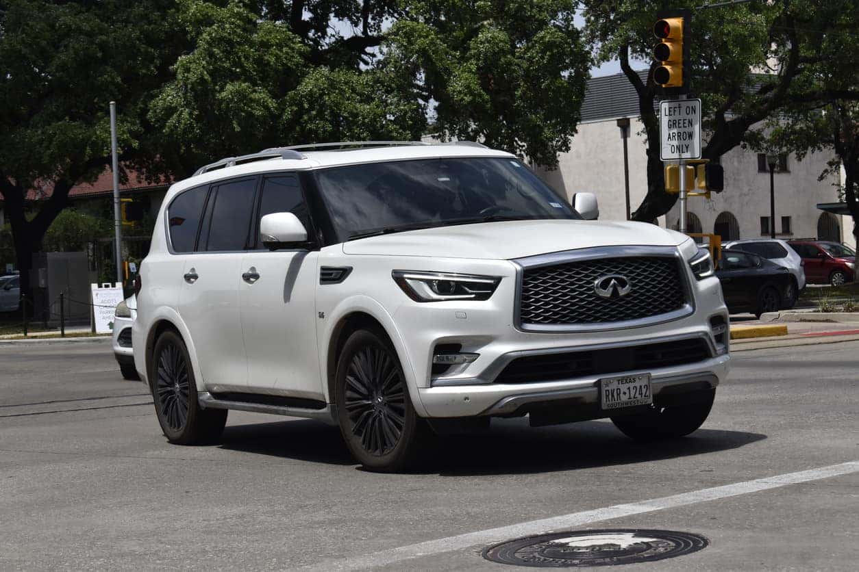 A portrait of a white Infinity QX80 SUV cruising in a downtown district