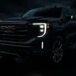 A black GMC Sierra Denali is parked on a dark, wet surface under moody skies, with its LED headlights and signature grille dramatically illuminated. The close-up angle emphasizes the truck's bold design and luxury detailing, perfectly representing the commanding presence of the GMC Sierra Denali.