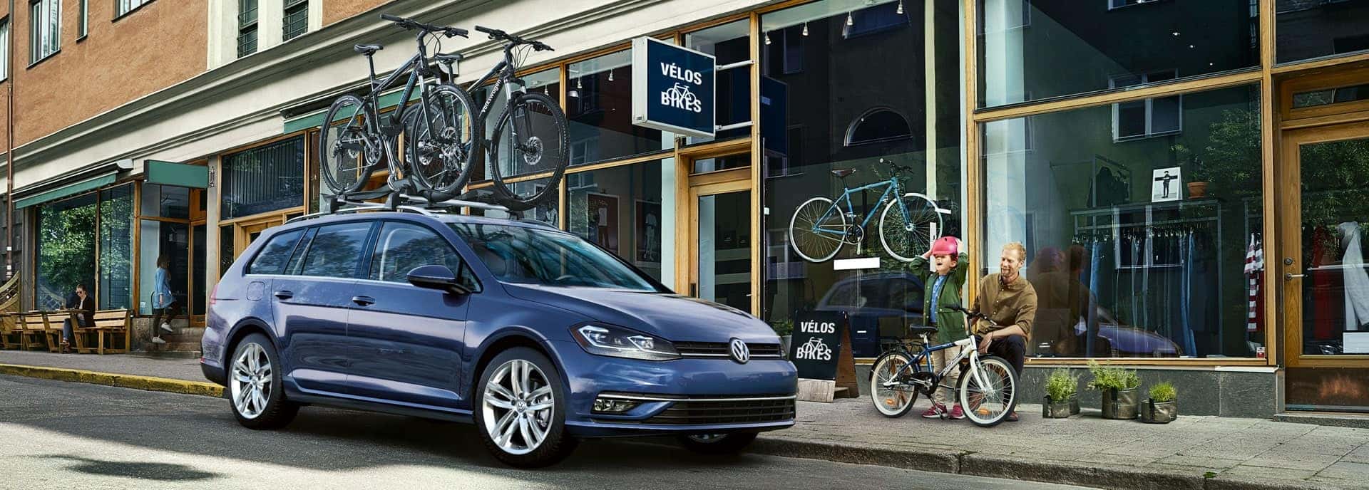 Blue 2019 Golf SportWagen at the bike shop. A little girl with a pink bicycle helmet is overjoyed. A man, presumably her father, seems happy but restrained.